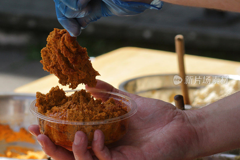 街头小吃市场的图片，不同种类的鹰嘴豆泥/橙色的番茄鹰嘴豆泥被盛在带有勺子的容器和罐子里/蓝色的食品卫生一次性塑料手套和手，希腊美食鹰嘴豆泥/鹰嘴豆泥，橄榄油，芝麻酱